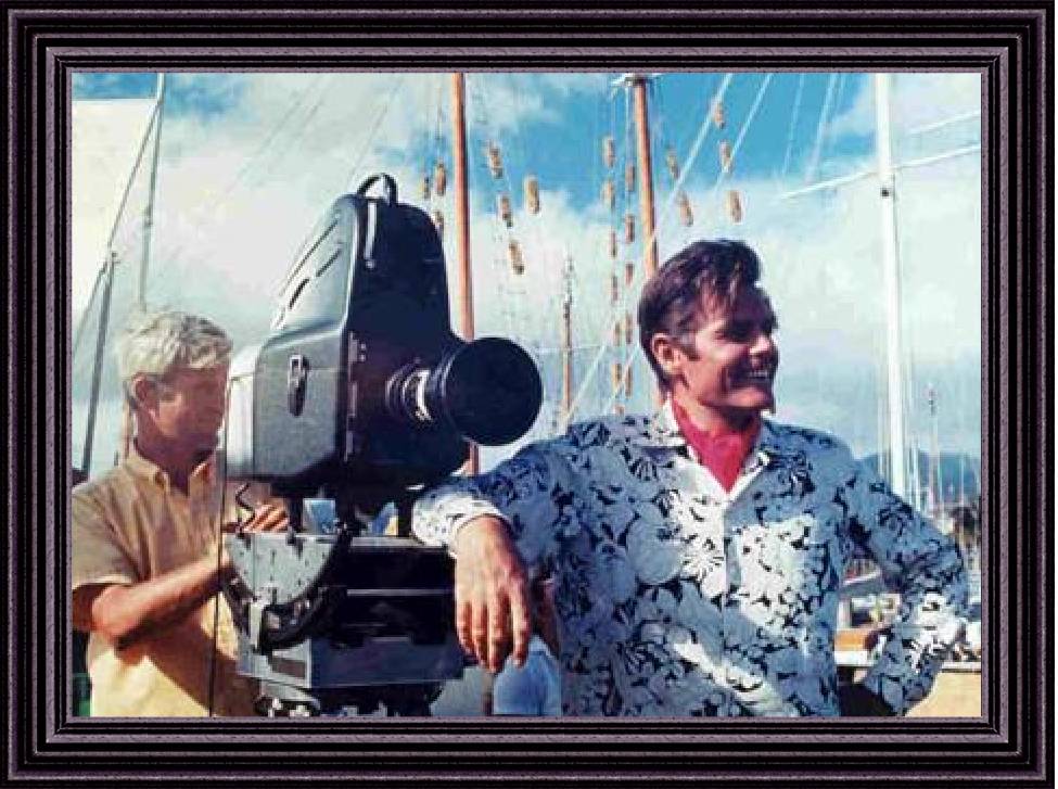 Jack Lord wering a black and white aloha shirt leaning on a television camera.Behind him is an unidentified cameraman. Was taken at the Ala Wai Yacht Harbor